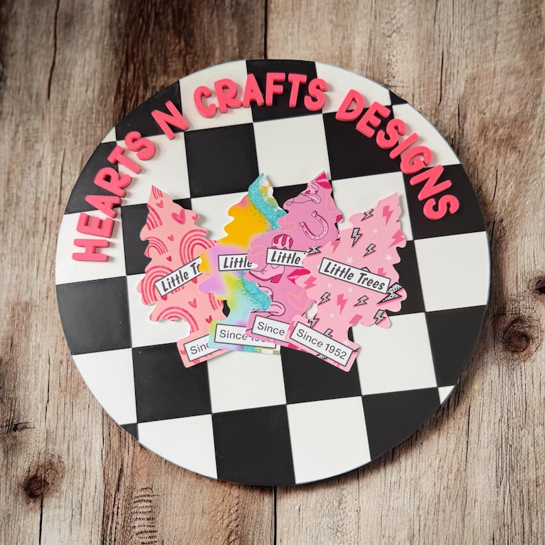 May include: A round, black and white checkered sign with the text "HEARTS N CRAFTS DESIGNS" in pink. The sign features pink tree-shaped cutouts with the words "Little Trees" and "Since 1952".