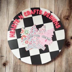 May include: A round, black and white checkered decorative piece with the text "HEARTS N CRAFTS DESIGNS" in pink. Three Christmas tree-shaped cutouts with the text "Little Trees" and "Since 1952" are on the surface.