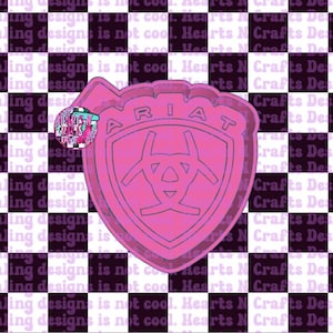 May include: A pink Ariat logo-shaped cookie cutter. The cutter features the brand name and a central design. The background is a checkerboard pattern with the text "Stealing designs is not cool. Hearts N Crafts Designs."