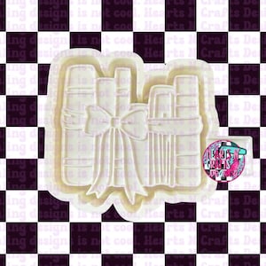 May include: A white plastic cookie cutter in the shape of a stack of books tied with a bow. The cutter has a decorative border and is set against a black and white checkered background.