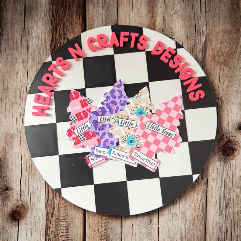 May include: A round decorative piece with a black and white checkered pattern. The words "HEARTS N CRAFTS DESIGNS" are in pink letters around the edge. Several tree-shaped cutouts with various patterns and the words "Little Trees" are on the surface.