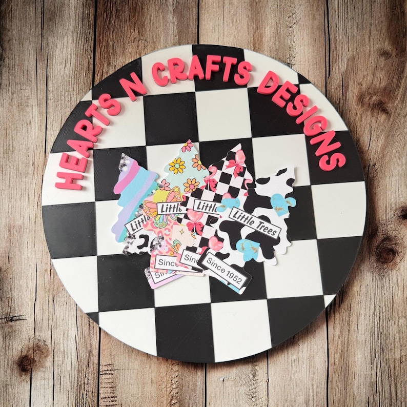 May include: A round, black and white checkered sign with the text "HEARTS N CRAFTS DESIGNS" in pink letters. Several tree-shaped cutouts with various patterns and the text "Little Trees" are attached to the sign.