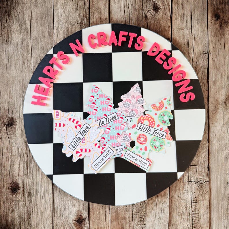 May include: A round, black and white checkered decorative piece with the text "HEARTS N CRAFTS DESIGNS" in pink. Three Christmas tree cutouts with the text "Little Trees" and "Since 1952" are on the piece.