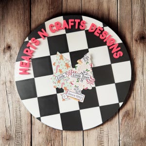May include: A round, black and white checkered sign with the text "HEARTS N CRAFTS DESIGNS" in pink letters. Two Christmas tree cutouts are on the sign, with the text "Little Trees" and "Since 1952".