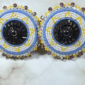 Peut inclure: Une paire de boucles d'oreilles en perles avec un motif circulaire. Les boucles d'oreilles sont blanches, bleues et jaunes avec un centre noir et un accent argenté. Les boucles d'oreilles sont entourées de cristaux transparents.