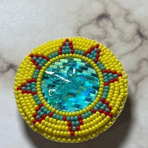 May include: A round beaded ornament featuring a turquoise hummingbird design in the centre. The ornament is surrounded by yellow beads with red and turquoise accents.