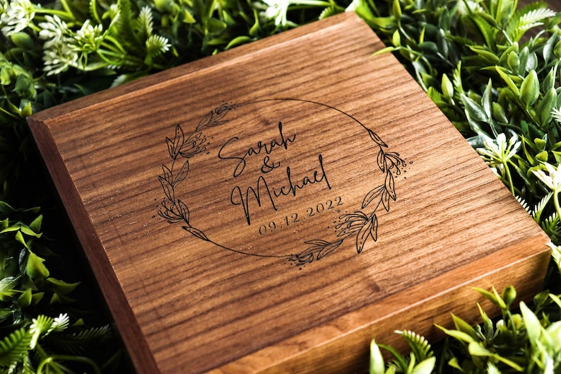 Op de afbeelding: Een houten doos met een gegraveerd ontwerp van een bloemenkrans op de deksel. De krans omsluit de tekst "Sarah & Michael" en de datum "09.12.2022".