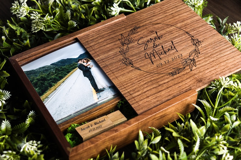 Op de afbeelding: Een houten doos met een schuifdeksel, met daarin een foto van een stel dat elkaar kust op een weg en een USB-stick. Het deksel is gegraveerd met de tekst "Sarah & Michael 09.12.2022" in een bloemkrans.