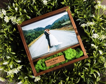 May include: A wooden framed photo of a couple embracing on a road, with green moss and a wooden USB drive. The USB drive is engraved with the names "Sarah & Michael" and the date "09.12.2022". The frame is surrounded by greenery.