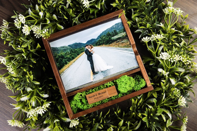 Op de afbeelding: Een houten doos met een glazen foto-inzet van een stel dat elkaar kust op een weg. De doos is bekleed met groen mos en heeft een klein houten plaatje met de tekst "Sarah & Michael 09.12.2022".