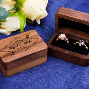May include: Two wooden ring boxes. One box is closed and engraved with the text "Sajjad & Alisha 10.12.2021". The other box is open and contains two rings. One ring has a clear stone and the other ring has a purple stone.