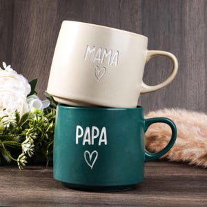 May include: Two ceramic mugs stacked. The top mug is cream-coloured with the word "MAMA" and a heart etched on it. The bottom mug is teal with the word "PAPA" and a heart etched on it. Both mugs have a rounded handle.