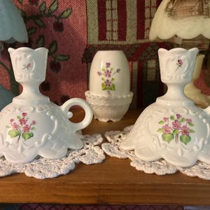 May include: Two white ceramic candle holders with a floral design. Each holder has a handle and is decorated with pink and green flowers. They sit on white lace doilies.