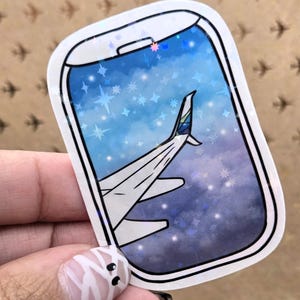 May include: A sticker featuring a stylized airplane wing viewed through a window against a starry sky. The window frame is white with a black outline. The sky transitions from blue to purple, with white stars.