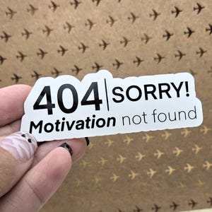 May include: A white sticker with black text that reads "404 SORRY! Motivation not found." The sticker is held against a brown background with a pattern of small airplane silhouettes.