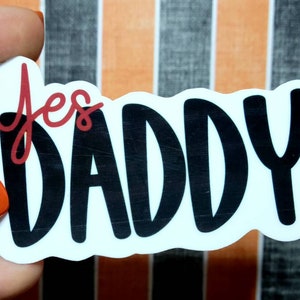 May include: A white sticker with the words "Yes DADDY" in black and red lettering. The sticker has a rounded rectangular shape. The background is a striped pattern of black, white, and orange.
