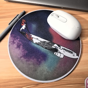 Peut inclure: Tapis de souris rond avec un motif d'avion stylisé et le mot "HAWAIIAN". Le tapis de souris a un fond de galaxie coloré, une souris sans fil blanche et un stylo gris sur une surface en bois.