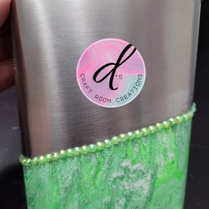 May include: A stainless steel flask with a green and white marbled design and a silver lid. The flask has a silver band with rhinestones around the top of the marbled design. A pink circle sticker with the text "D's Craft Room Creations" is on the front of the flask.