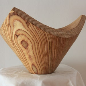 May include: A wooden sculpture with a unique, organic shape. The sculpture is made of light brown wood with visible grain patterns. It is displayed on a white fabric surface.