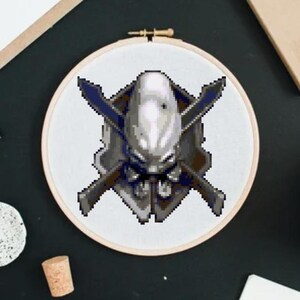 Könnte beinhalten: Ein weißes und graues Stickmuster eines Helms mit gekreuzten Schwertern auf einem weißen Stoffhintergrund. Das Design befindet sich in einem runden Stickrahmen.