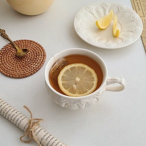 May include: A white teacup with a lemon slice in it, sitting on a white saucer. The teacup has a decorative design and a handle. The saucer has a floral design. There is a wicker coaster and a gold spoon in the background.