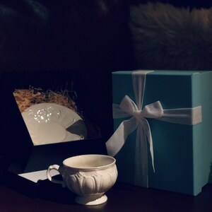 May include: A white porcelain teacup and saucer with a decorative floral design, next to a wrapped gift box with a light blue lid and a white satin ribbon.