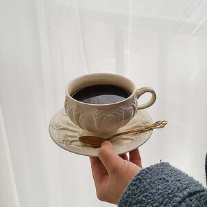 May include: A white ceramic coffee cup with intricate floral designs, filled with black coffee, sits on a matching saucer with a gold spoon.