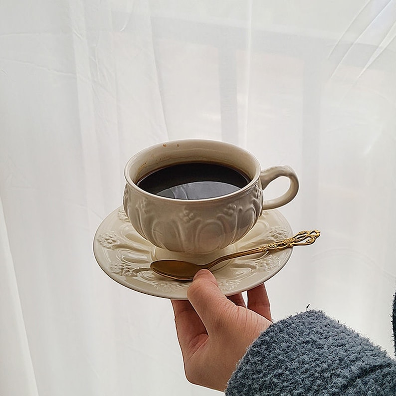 May include: A white ceramic coffee cup with intricate floral designs, filled with black coffee, sits on a matching saucer with a gold spoon.