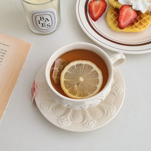 May include: A white teacup with a lemon slice in a cup of tea. The teacup is on a white saucer with a floral design. The teacup has a handle and is filled with tea.
