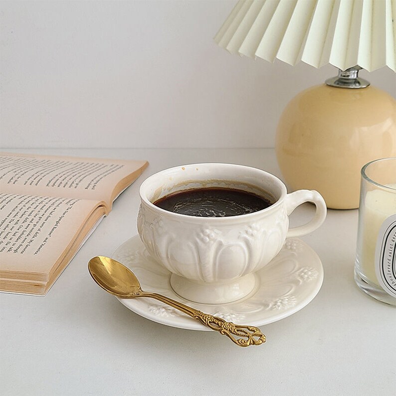 May include: A white ceramic coffee cup with intricate floral designs, filled with dark coffee, sits on a white saucer with a gold spoon. The cup is on a white surface next to a book and a yellow lamp.