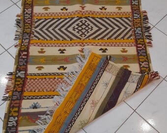 Puede incluir: Una alfombra tejida a mano colorida con patrones geométricos en tonos de marrón, amarillo, rojo y azul. La alfombra tiene un borde de flecos y está doblada por la mitad.