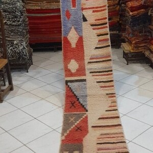 May include: A long, rectangular rug with a geometric pattern in shades of red, orange, brown, blue, and white. The rug has a fringe on the bottom.