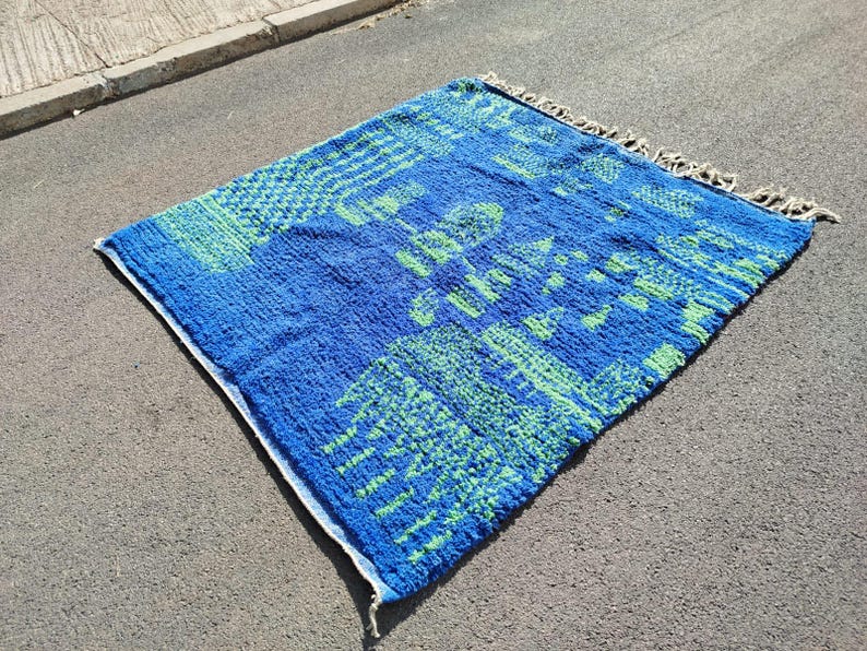 May include: A blue and green patterned rug with a fringed border. The rug features a geometric design with a variety of shapes and sizes.