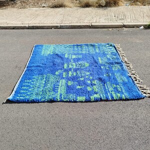 May include: A blue and green patterned rug with fringe on the edges. The rug is laid out on a grey asphalt surface.