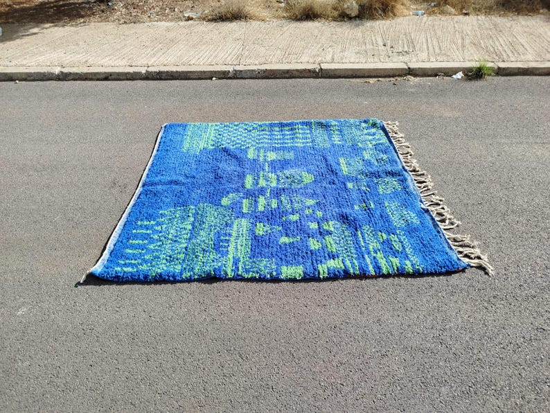 May include: A blue and green patterned rug with fringe on the edges. The rug is laid out on a grey asphalt surface.