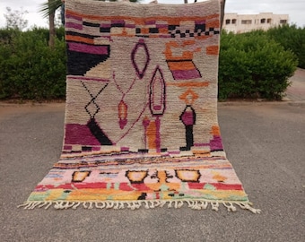 May include: A rectangular, hand-woven rug with a beige background and colorful geometric patterns. The design includes black, purple, orange, and pink shapes. The rug has a fringed edge and is displayed outdoors.