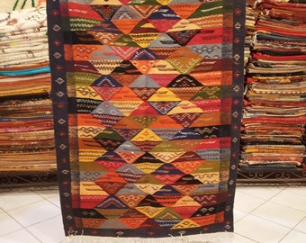 May include: A colorful, geometric patterned rug with a dark blue border. The rug is made of woven wool and has a fringe on the bottom.