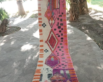 Puede incluir: Una alfombra larga y estrecha con un patrón geométrico en tonos de rosa, naranja y morado. La alfombra tiene flecos en el borde inferior.