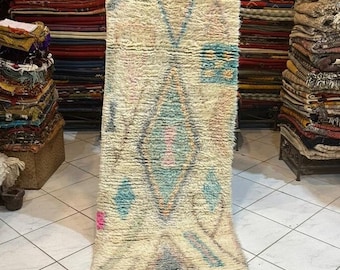 May include: A white Moroccan rug with a geometric pattern in shades of blue, pink, and beige. The rug has a fringe border and is displayed in a shop.