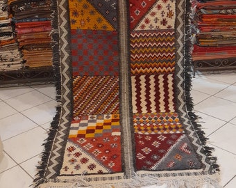Puede incluir: Una alfombra marroquí colorida con un patrón geométrico en tonos de rojo, amarillo, negro y blanco. La alfombra está doblada por la mitad y tiene flecos en los bordes.