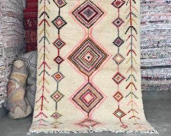 May include: A white rug with a geometric pattern in shades of pink, red, black, and gray. The rug has a fringe border and the text "2,18/1,38" is printed on the bottom.