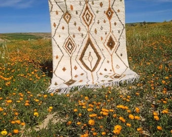 May include: A white Moroccan rug with a diamond pattern in shades of brown, green, and orange. The rug is laid out in a field of yellow flowers.