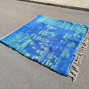 May include: A blue and green patterned rug with white fringe. The rug has a geometric design with a Moroccan style.
