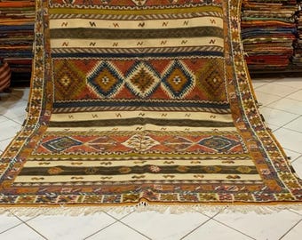 May include: A large, rectangular rug with a cream-colored background and intricate geometric patterns in shades of brown, orange, blue, and green. The rug features diamond and stripe designs, with a fringed edge. The rug is displayed on a tiled floor.