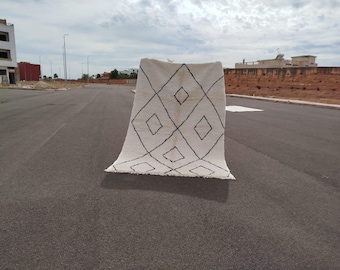 May include: A cream-colored rug with a geometric diamond pattern in black. The rug is displayed outdoors on a paved surface, showcasing its design and texture. The rug's fringe is visible at the bottom.