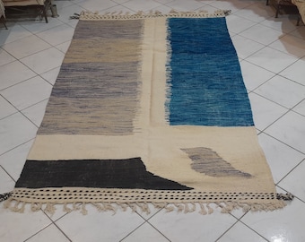 Op de afbeelding: Een handgeweven tapijt met een geometrisch ontwerp in tinten blauw, grijs, zwart en wit. Het tapijt heeft een franje rand.