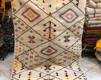 May include: A white Moroccan rug with a geometric pattern in shades of red, blue, yellow, green, and pink. The rug has a fringe border and is made of wool.