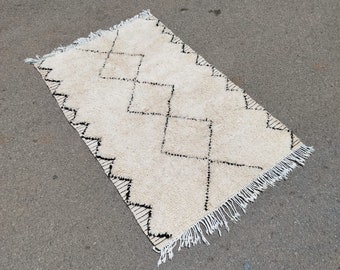 May include: A white and black Moroccan rug with a diamond pattern and fringe. The rug is laid out on a gray asphalt surface.