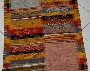 May include: A colorful Moroccan rug with a geometric pattern in shades of red, yellow, blue, and black. The rug has a fringe border and is made of wool.