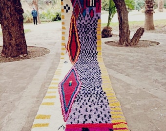 May include: A long, white rug with a colourful geometric pattern. The rug has a diamond shape in the centre with yellow, red, blue, and green accents. The rug is made of wool and has a fringe on the ends.